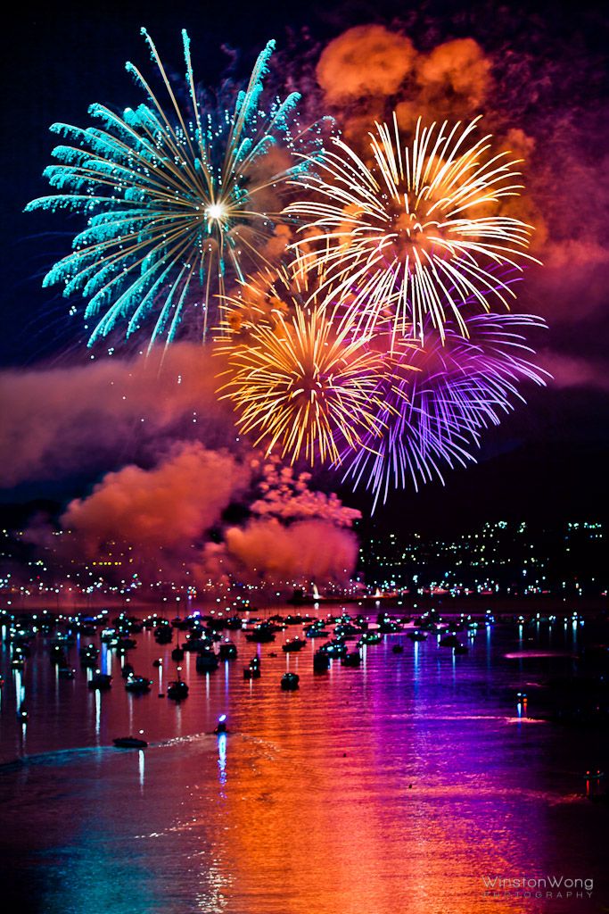 fireworks are lit up in the night sky over water and buildings with lights on them