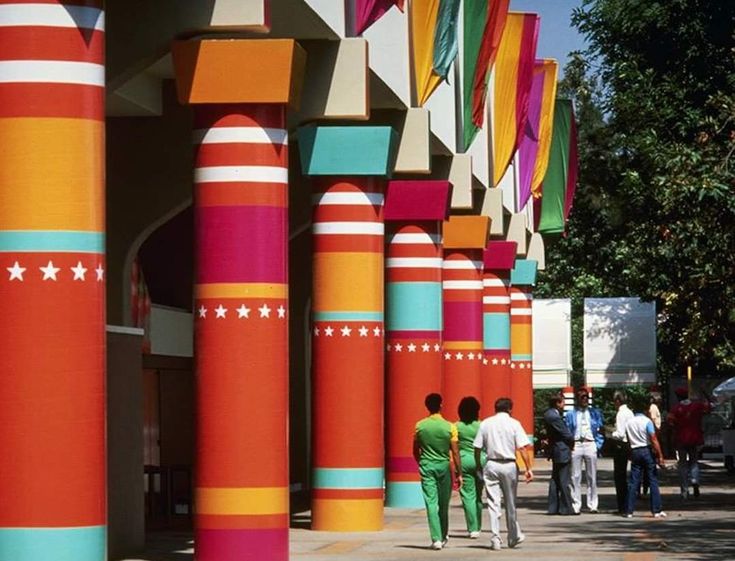 people walking past brightly colored columns on the side of a building with trees in the background