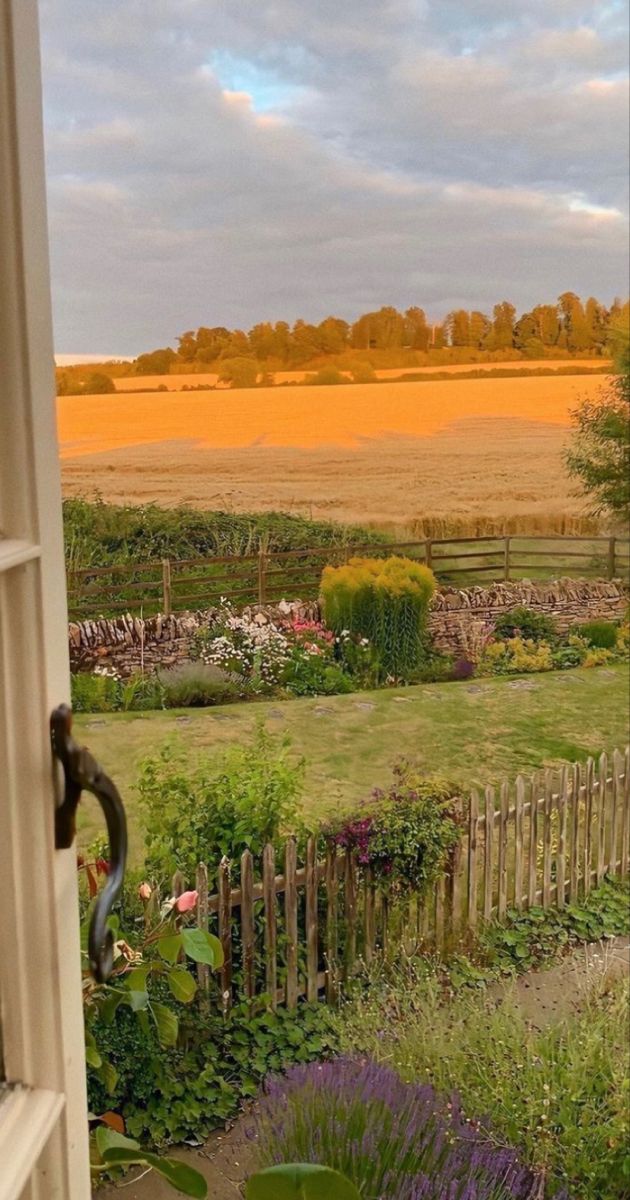 an open window looking out onto a garden and field in the distance with trees, shrubs, and flowers