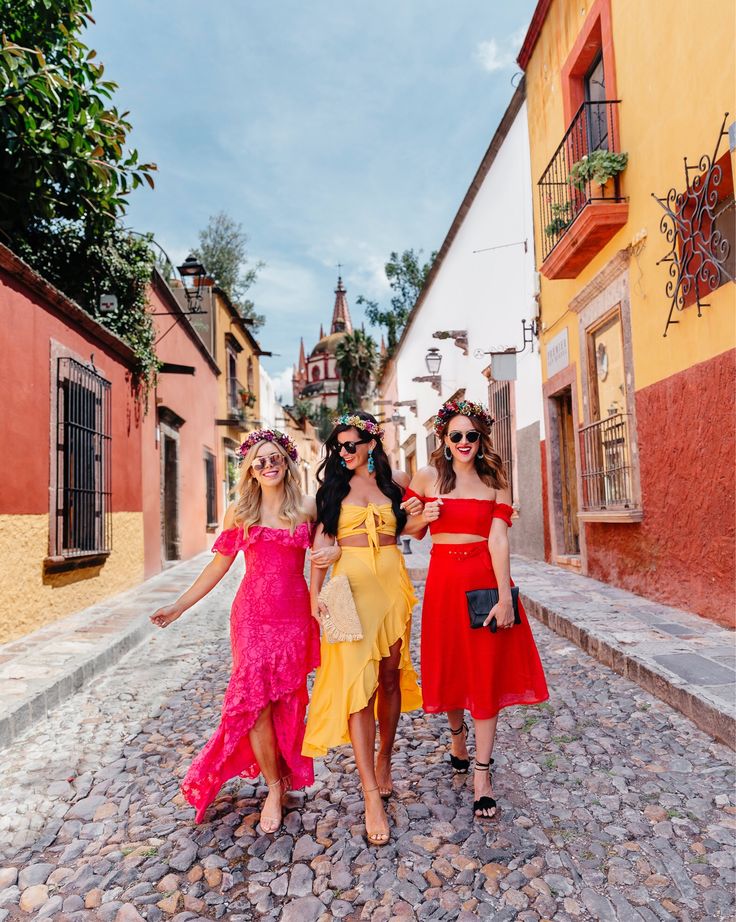 three women in colorful dresses walking down a cobblestone street with their arms around each other