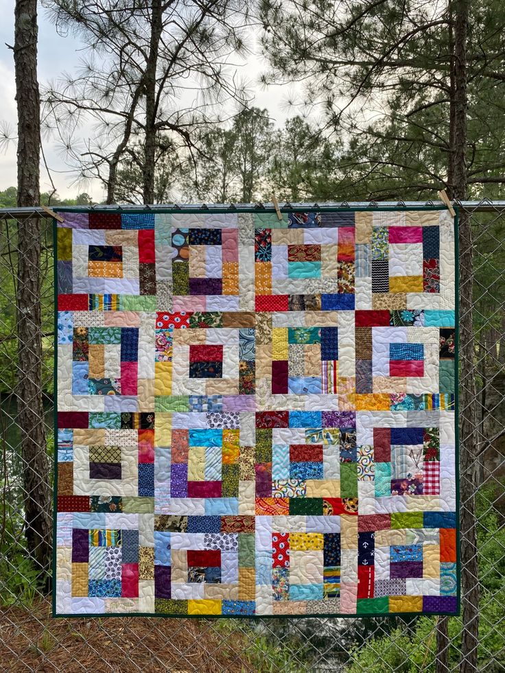 a quilt hanging on a chain link fence with trees in the backgroung