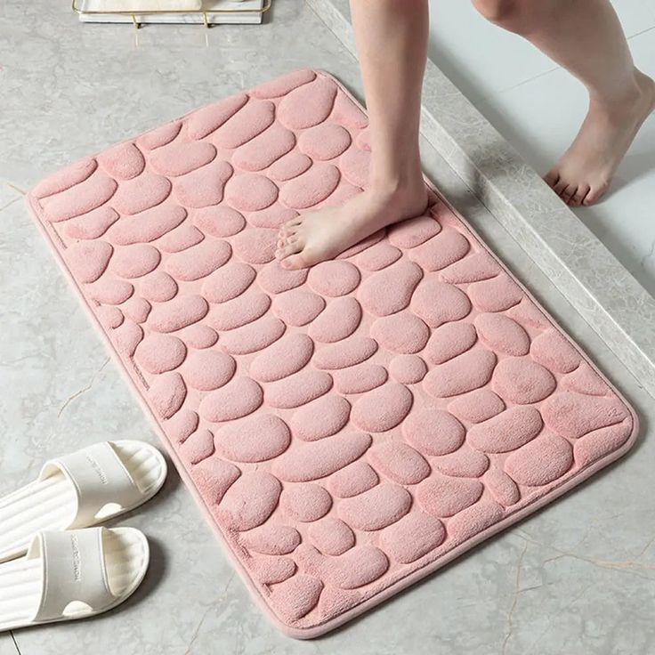 a person standing on a bathroom rug next to a pair of white slippers and sandals