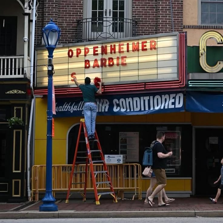 a man is standing on a ladder in front of a movie theater while people walk by