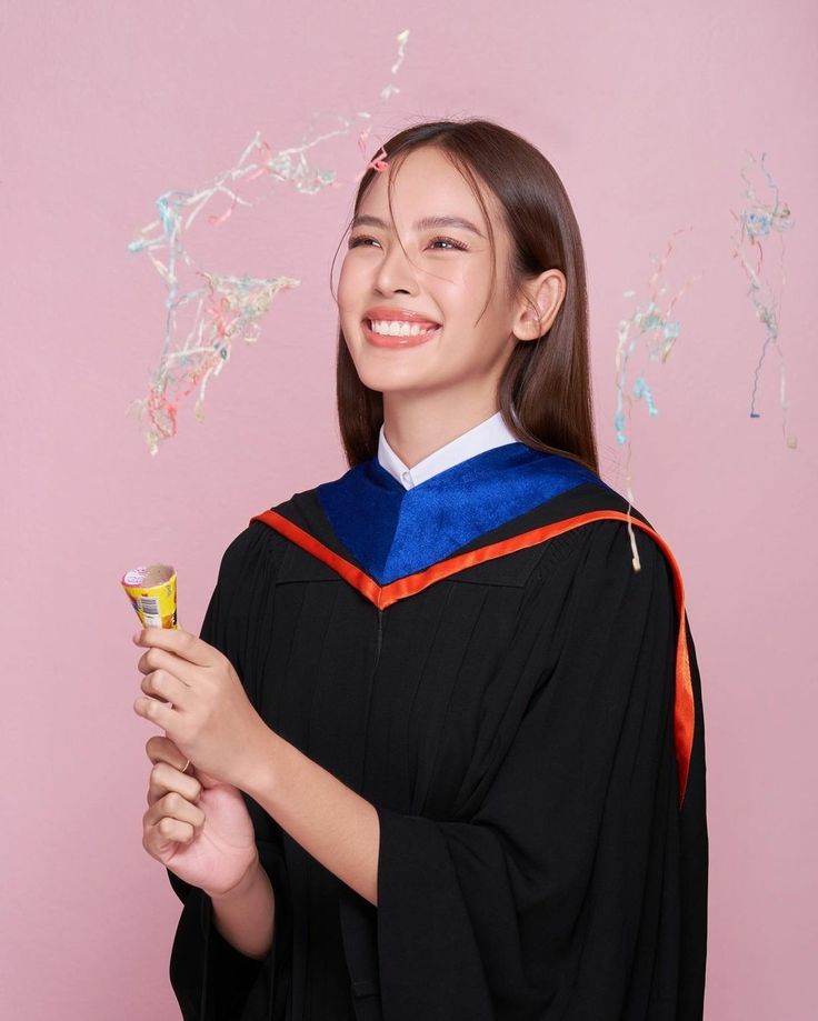 a young woman in graduation gown holding a small cup and smiling at the camera while standing against a pink background