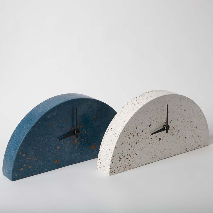 two clocks made out of concrete sitting side by side on a white surface, one is blue and the other is grey