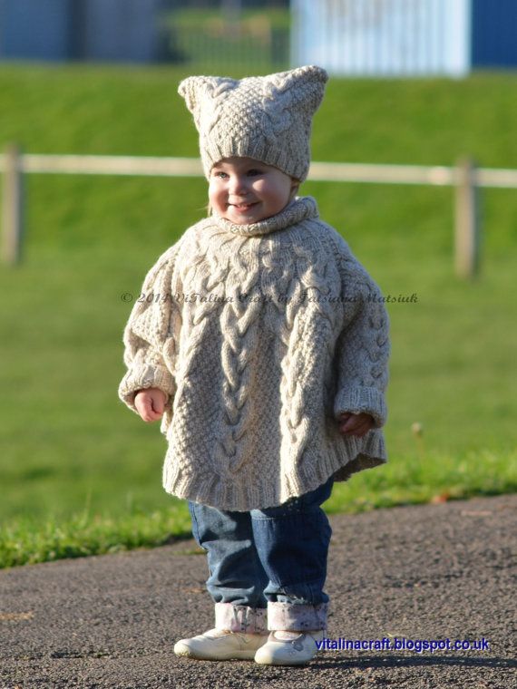 a little boy wearing a knitted sweater and hat standing on the side of a road