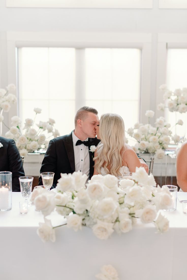 a bride and groom kissing in front of their guests at a wedding reception with white flowers on the table