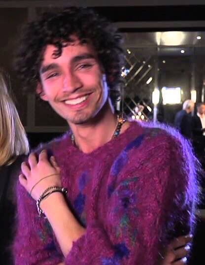 a man with curly hair wearing a purple sweater smiles at the camera while standing next to a woman