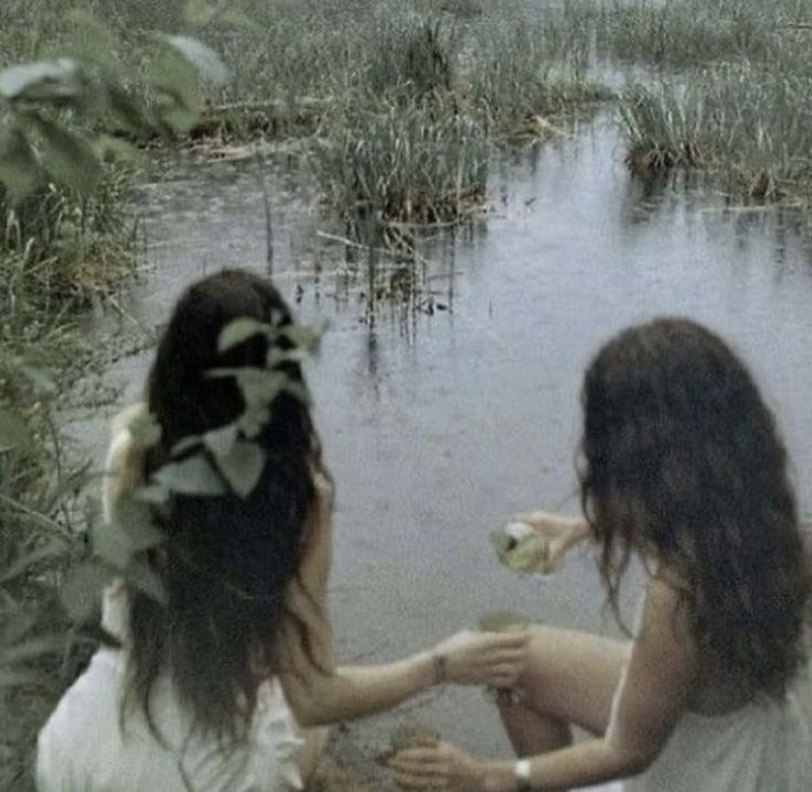 two young women sitting on the ground in front of a body of water and plants