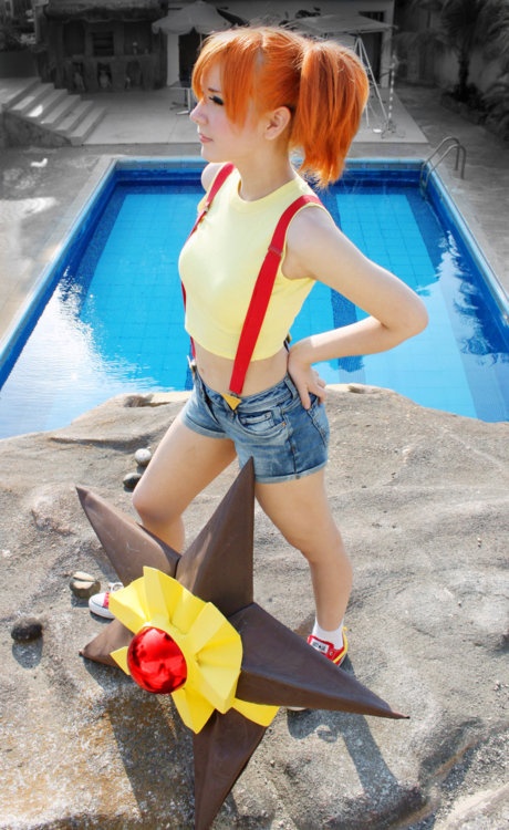 a woman with red hair standing in front of a pool wearing a yellow shirt and blue jean shorts