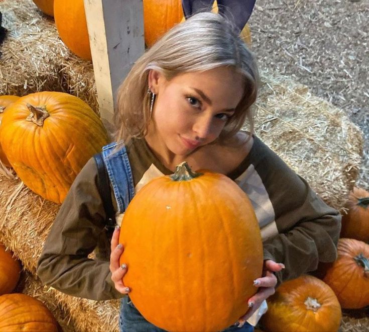 a girl holding a pumpkin in front of hay bales with other pumpkins behind her