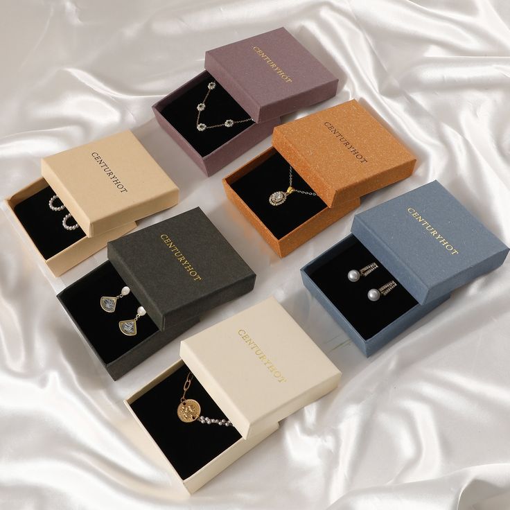 six different colored boxes with necklaces in them on a white cloth covered bed sheet