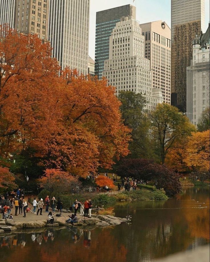 many people are walking around in the park near some water and trees with orange leaves