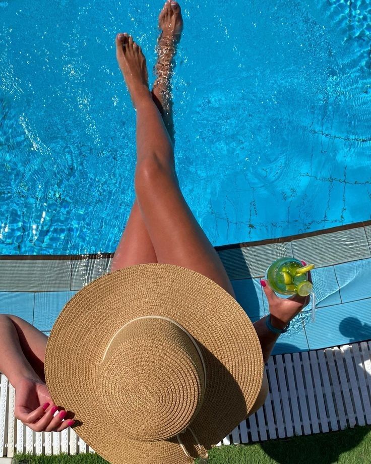 a woman in a straw hat sitting next to a swimming pool holding a drink and looking at the camera