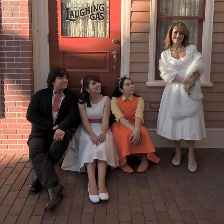 three women and two men sitting on the front steps of a house, one woman is wearing an orange dress