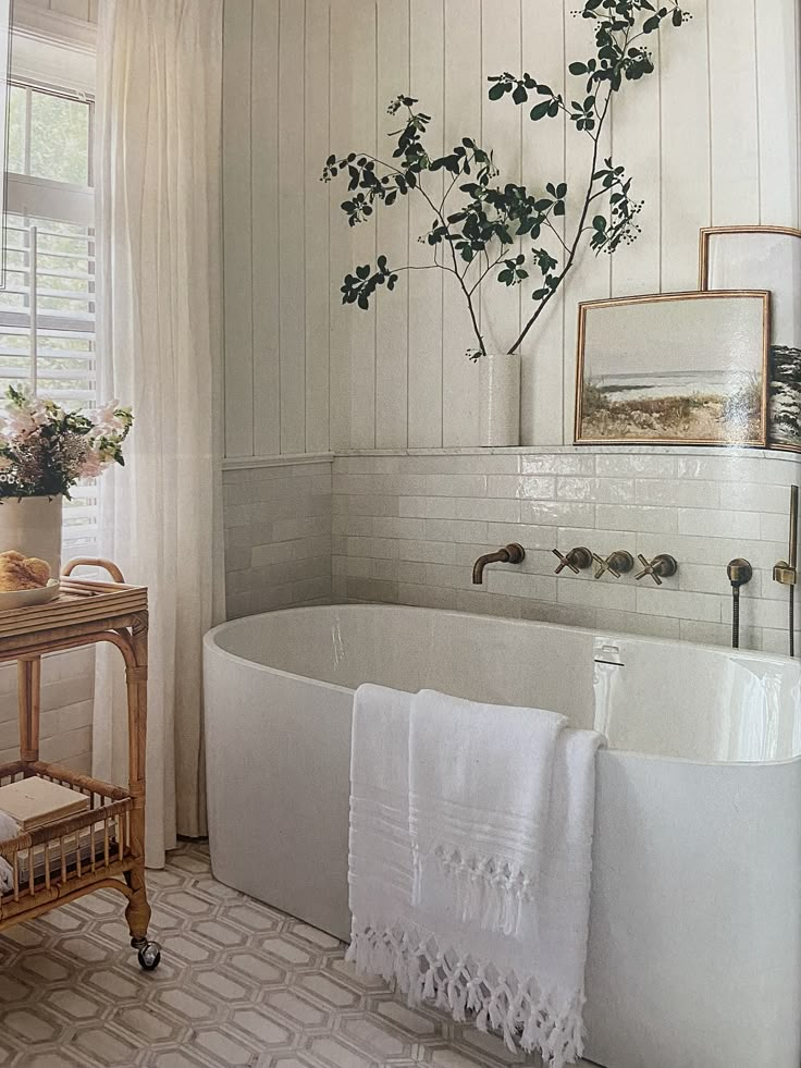 a white bath tub sitting under a window next to a wooden table with flowers on it