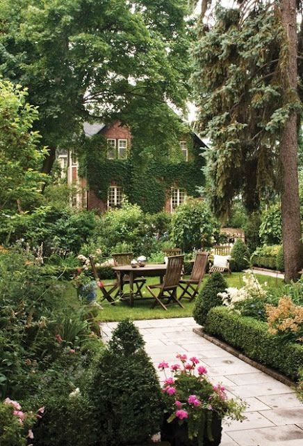 a table and chairs in the middle of a garden with trees, bushes and flowers