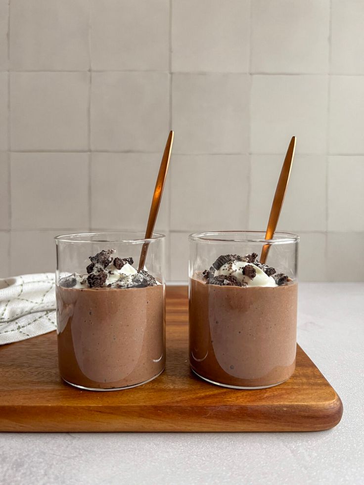 two glasses filled with chocolate pudding on top of a wooden cutting board next to a white tile wall