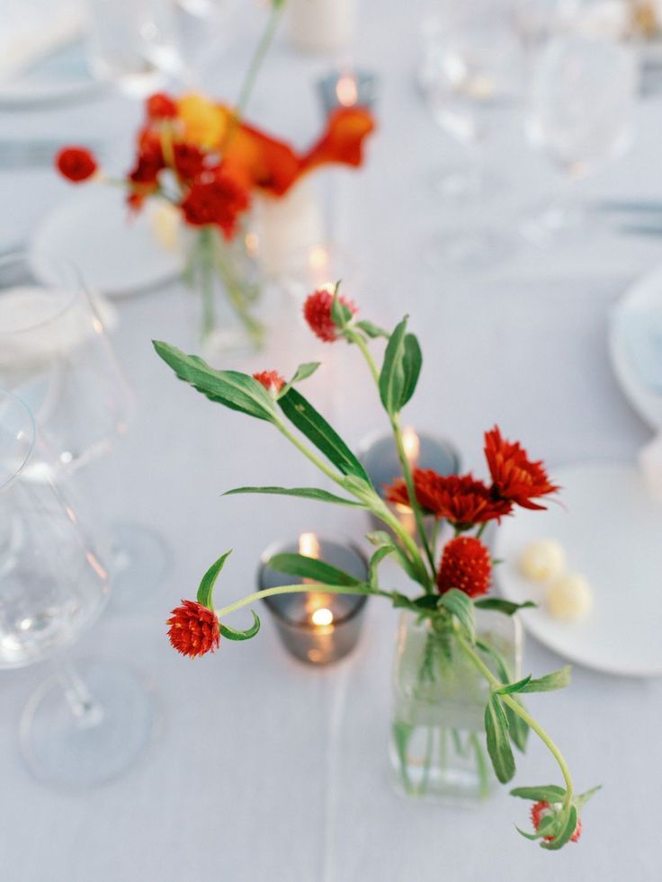 the table is set with white linens, silverware and red flowers in vases