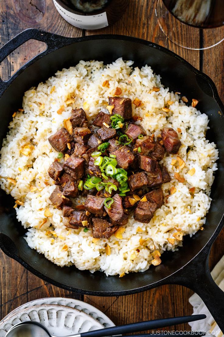 a skillet filled with rice and meat on top of a wooden table next to utensils