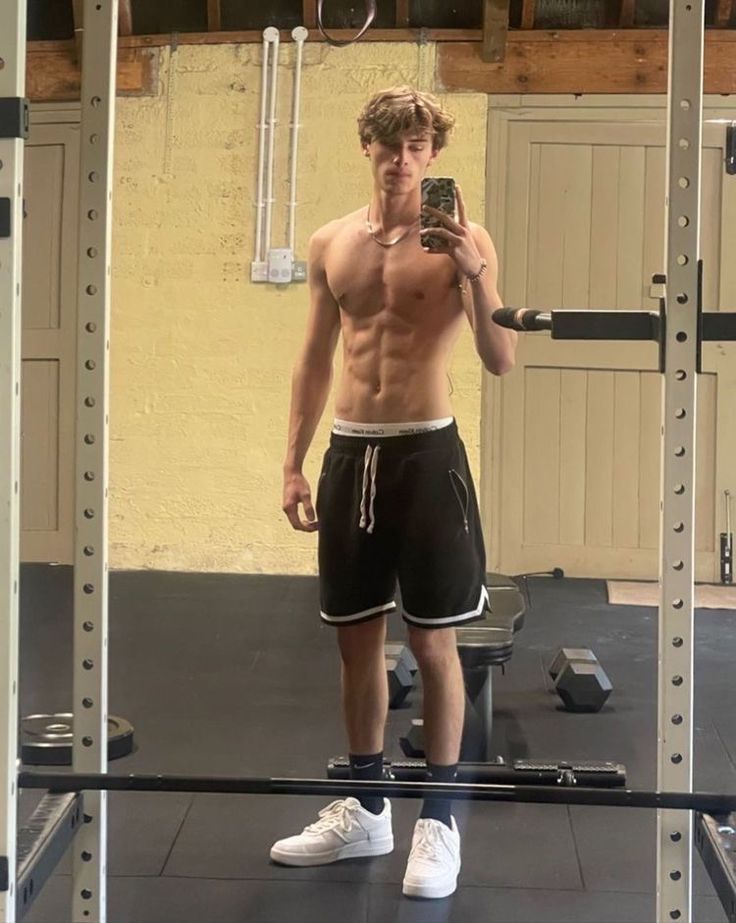 a shirtless man takes a selfie in the mirror while standing on a gym floor