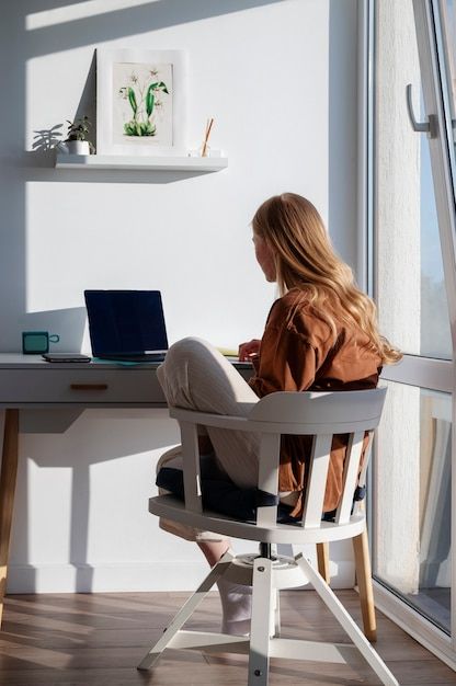 a woman sitting in a chair at a desk with a laptop on her lap looking out the window