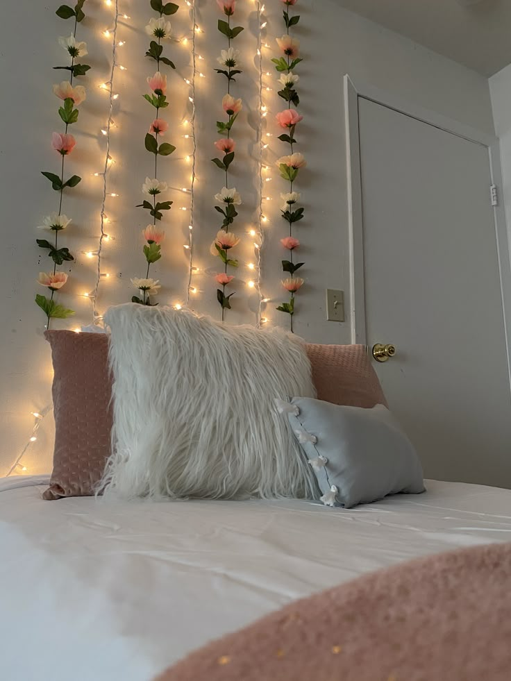 a white bed topped with lots of pillows and flowers next to a wall covered in lights