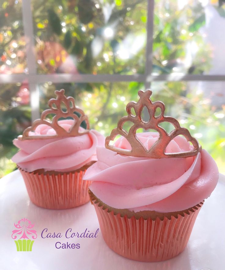 two cupcakes with pink frosting and gold crowns on top are sitting in front of a window