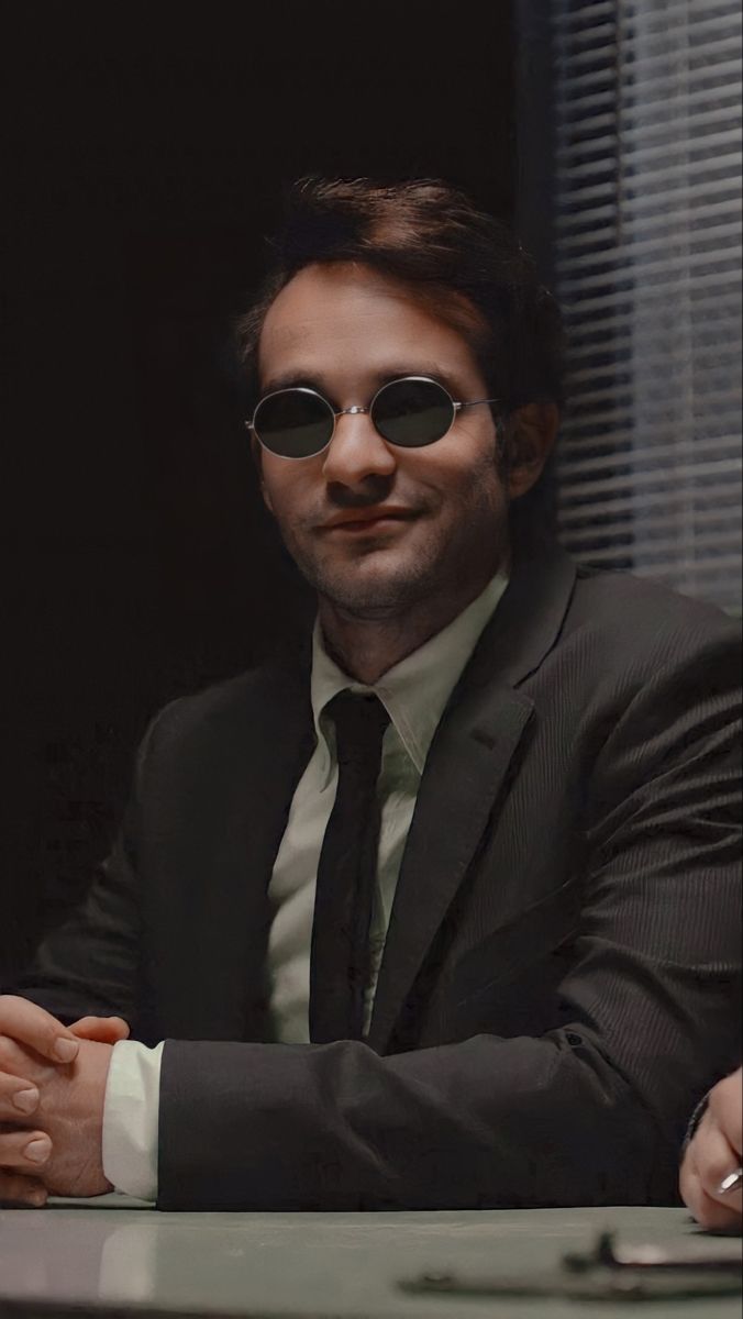 a man in a suit and sunglasses sitting at a table with his hands folded out