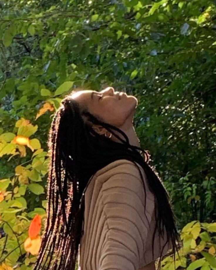 a woman with dreadlocks standing in front of some trees and bushes looking up at the sky