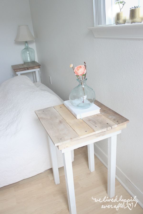 a small wooden table with a flower in a glass vase on it next to a bed