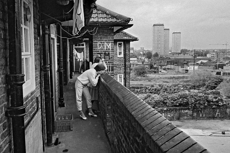 black and white photograph of man leaning against wall on balcony overlooking cityscape in the distance