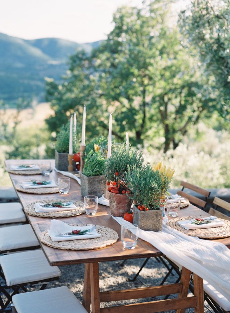 an outdoor table set with place settings and candles on it, surrounded by greenery