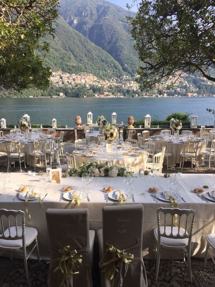 an outdoor table set up with white linens and flowers, overlooking the water's edge