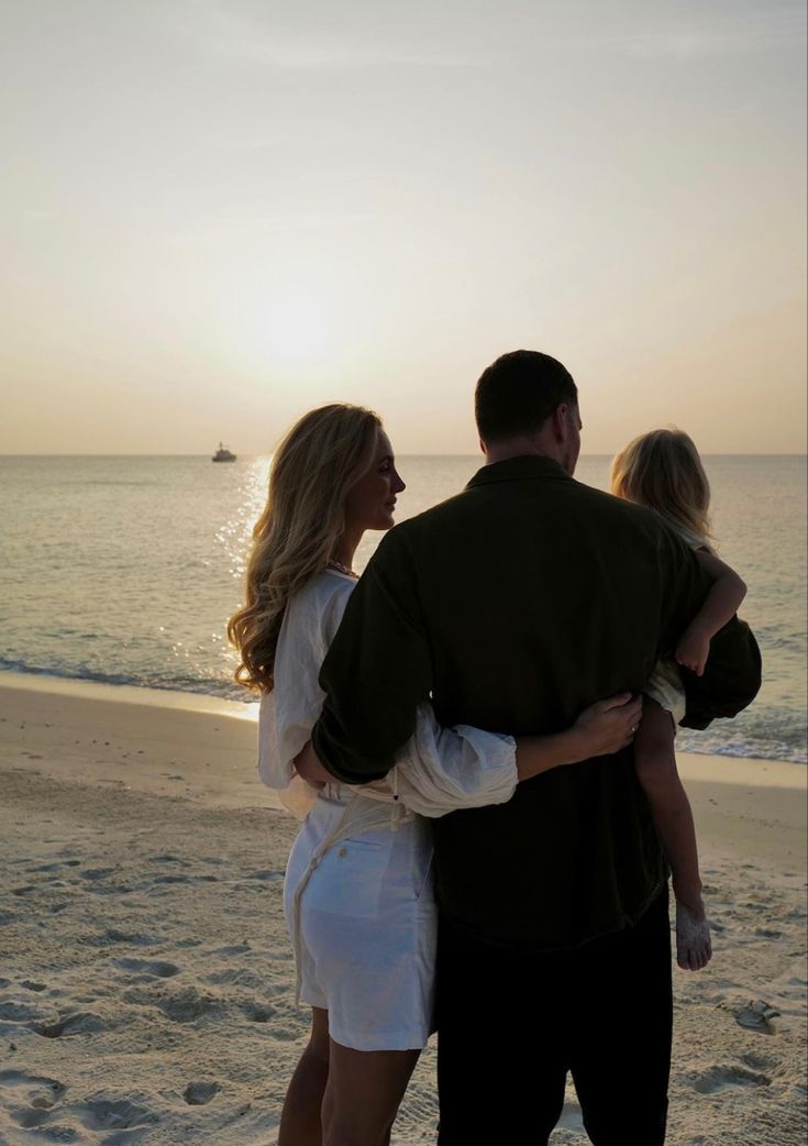 a man and woman standing on the beach with their arms around each other as the sun sets