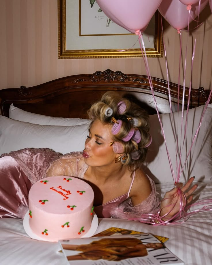 a woman laying in bed with a pink cake and balloons