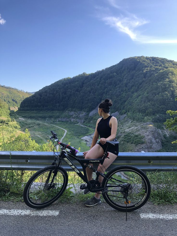 a woman sitting on top of a bike next to a lush green mountain side under a blue sky