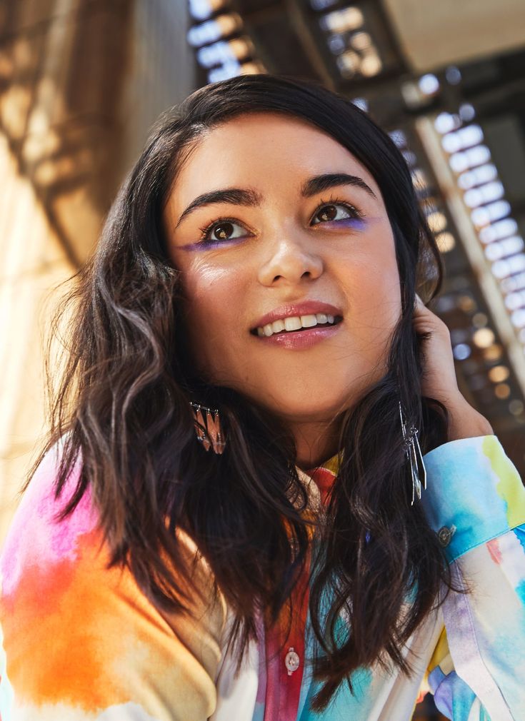 a woman with long dark hair and blue eyes smiles while holding her hand to her ear