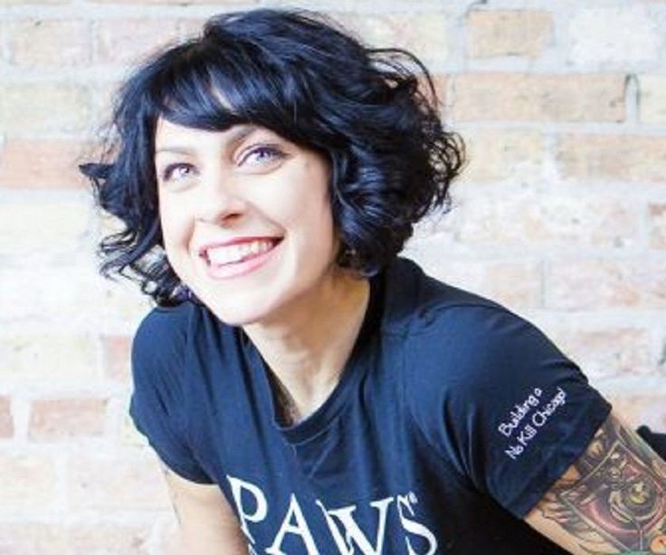 a woman with black hair and tattoos on her arm smiles at the camera while sitting in front of a brick wall