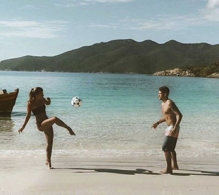 a man and woman playing soccer on the beach with a boat in the water behind them