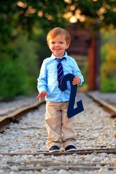 a young boy wearing a blue shirt and tie standing on train tracks with the number two