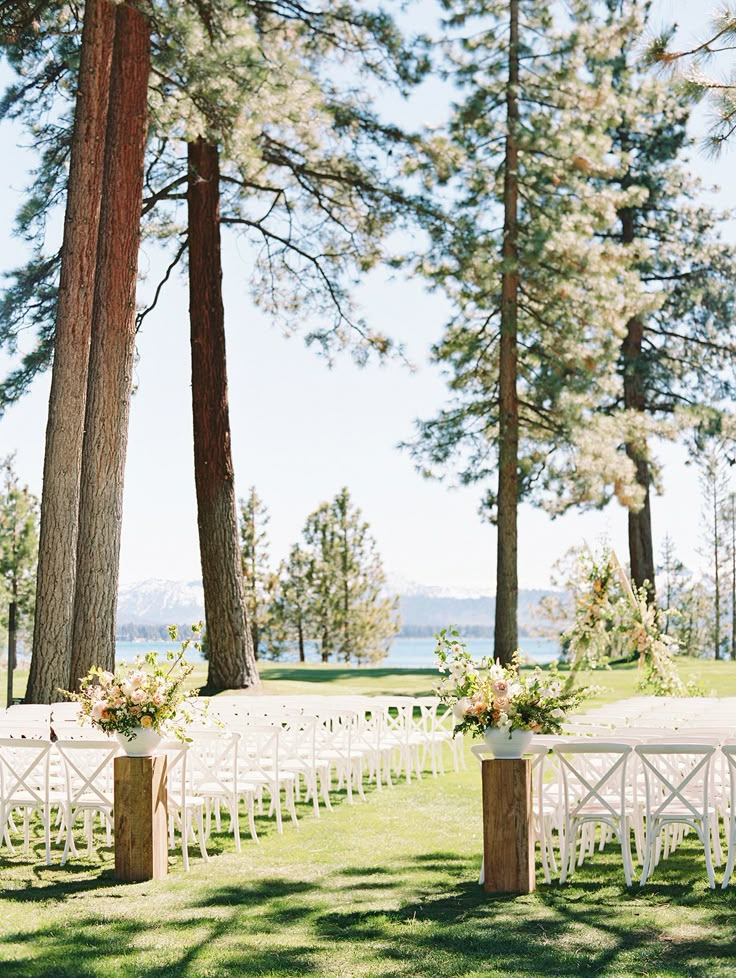 an outdoor ceremony setup with white chairs and floral centerpieces in front of pine trees