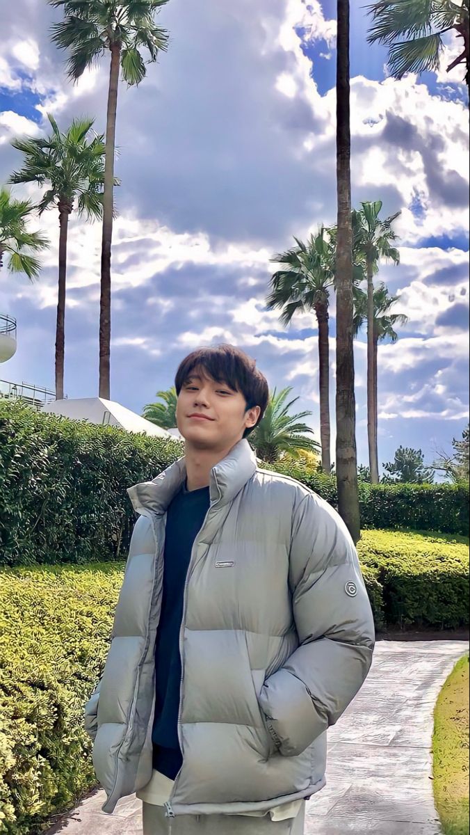 a man standing in front of palm trees wearing a puffy gray jacket and blue t - shirt