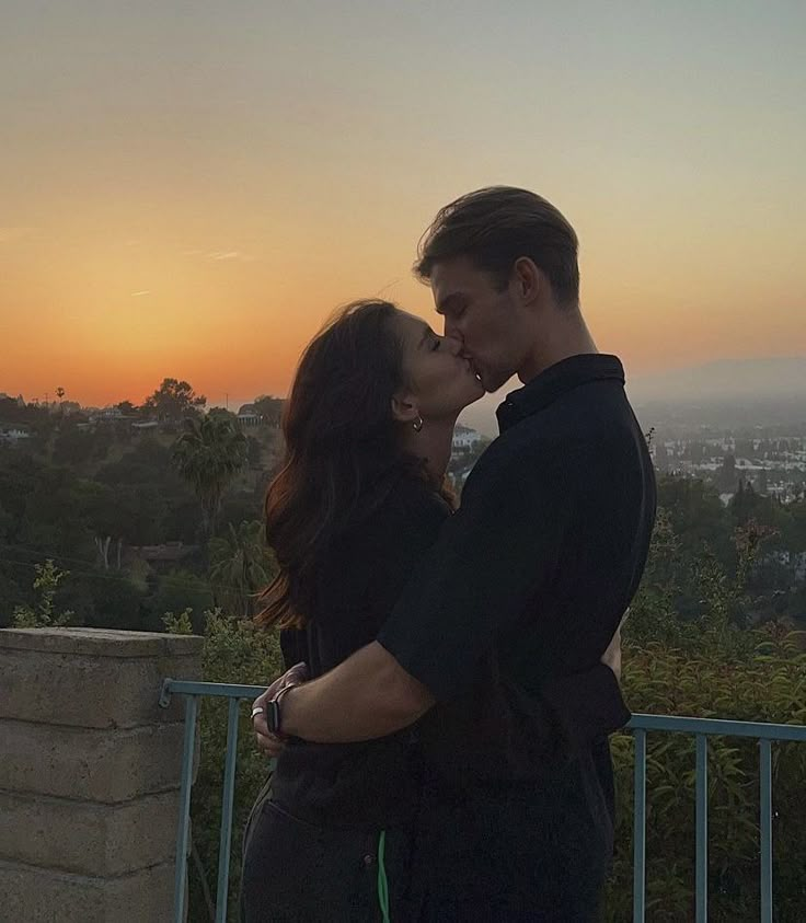 a man and woman standing next to each other near a fence with the sun setting in the background