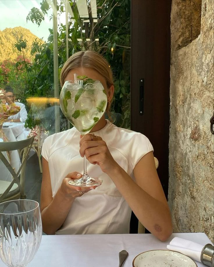 a woman sitting at a table holding a wine glass in front of her face with water inside