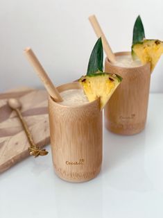 two glasses filled with pineapple drink on top of a white table next to a cutting board