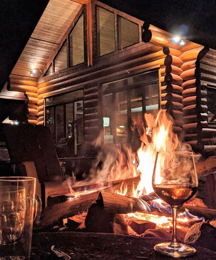 a glass of wine sitting on top of a fire pit in front of a log cabin