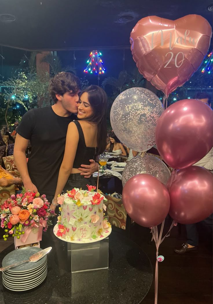 two people standing next to each other near a table with cake and balloons on it