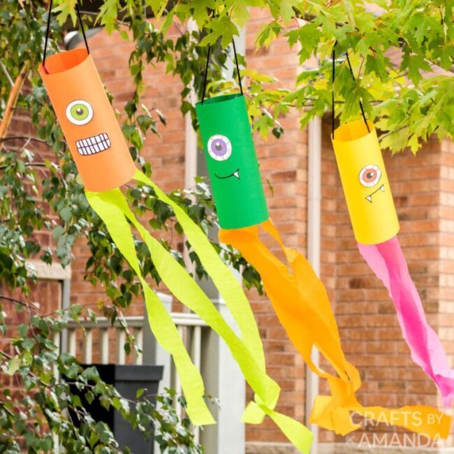 three colorful paper tubes hanging from a tree with googly eyes and mouth on them