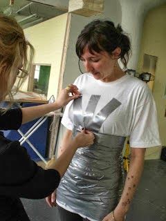 a woman is helping another woman put on her silver shirt in the room with other people